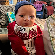 Jaxson is registered to the contest to win money with this photo: baby, infant, person, clothing, beanie, red_sleeves, striped_outfit, indoor, living_room, blanket, furniture, couch, basket, flooring, expression, face, headwear, portrait, newborn, holding