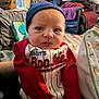 baby, infant, person, clothing, beanie, red_sleeves, striped_outfit, indoor, living_room, blanket, furniture, couch, basket, flooring, expression, face, headwear, portrait, newborn, holding