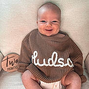 Hudson is registered to the contest to win money with this photo: baby, infant, smile, sweater, knitwear, diaper, chair, wooden_sign, age_marker, cute, happy, indoors, portrait, child, person, cozy, milestone, skin, head, legs
