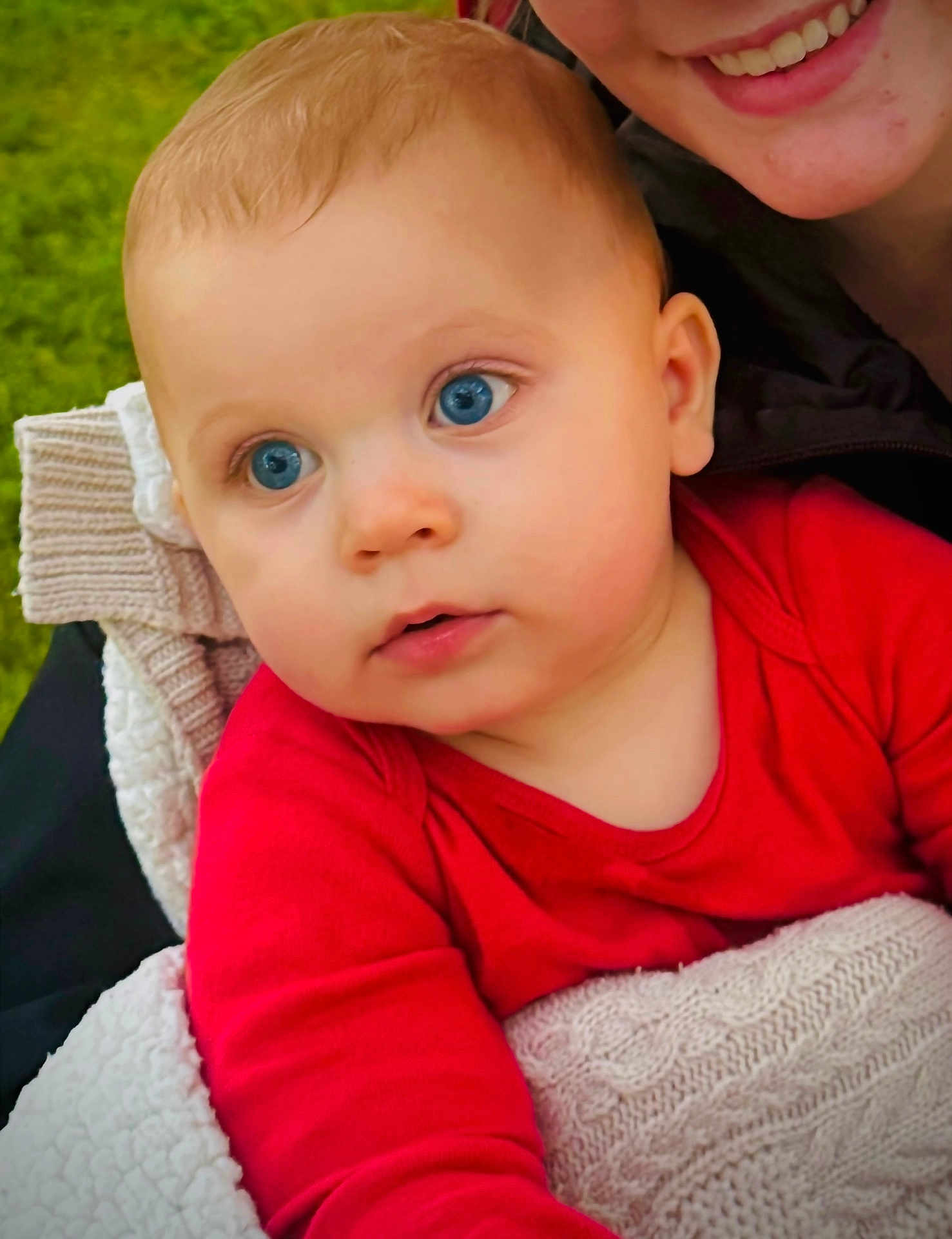 Warren is registered to the contest to win money with this photo: baby, child, blue_eyes, red_clothing, blanket, knit, face, smile, person, cuddle, portrait, outdoor, close_up, infant, young_child, happy, skin, head, warm, soft_texture