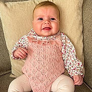Remy is registered to the contest to win money with this photo: baby, infant, pink_outfit, knitted_clothing, floral_shirt, couch, cushion, indoor, child, portrait, cute, smiling, seated, cozy, texture, furniture, small_hands, legs, head, face