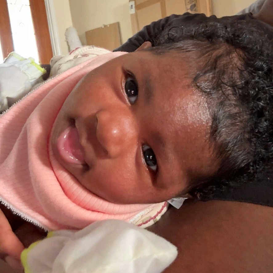 Kelsia is registered to the contest to win money with this photo: arm, art, baby, child, closeup, curly_hair, door, face, happy, indoor, infant, light, mittens, person, pink_bib, portrait, resting, skin, smiling, wall