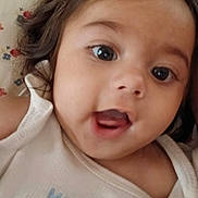 Selena is registered to the contest to win money with this photo: baby, infant, face, close_up, curly_hair, white_clothing, expression, mouth_open, eyes, skin, portrait, cute, child, young, person, lying_down, fabric, pattern, floral, home