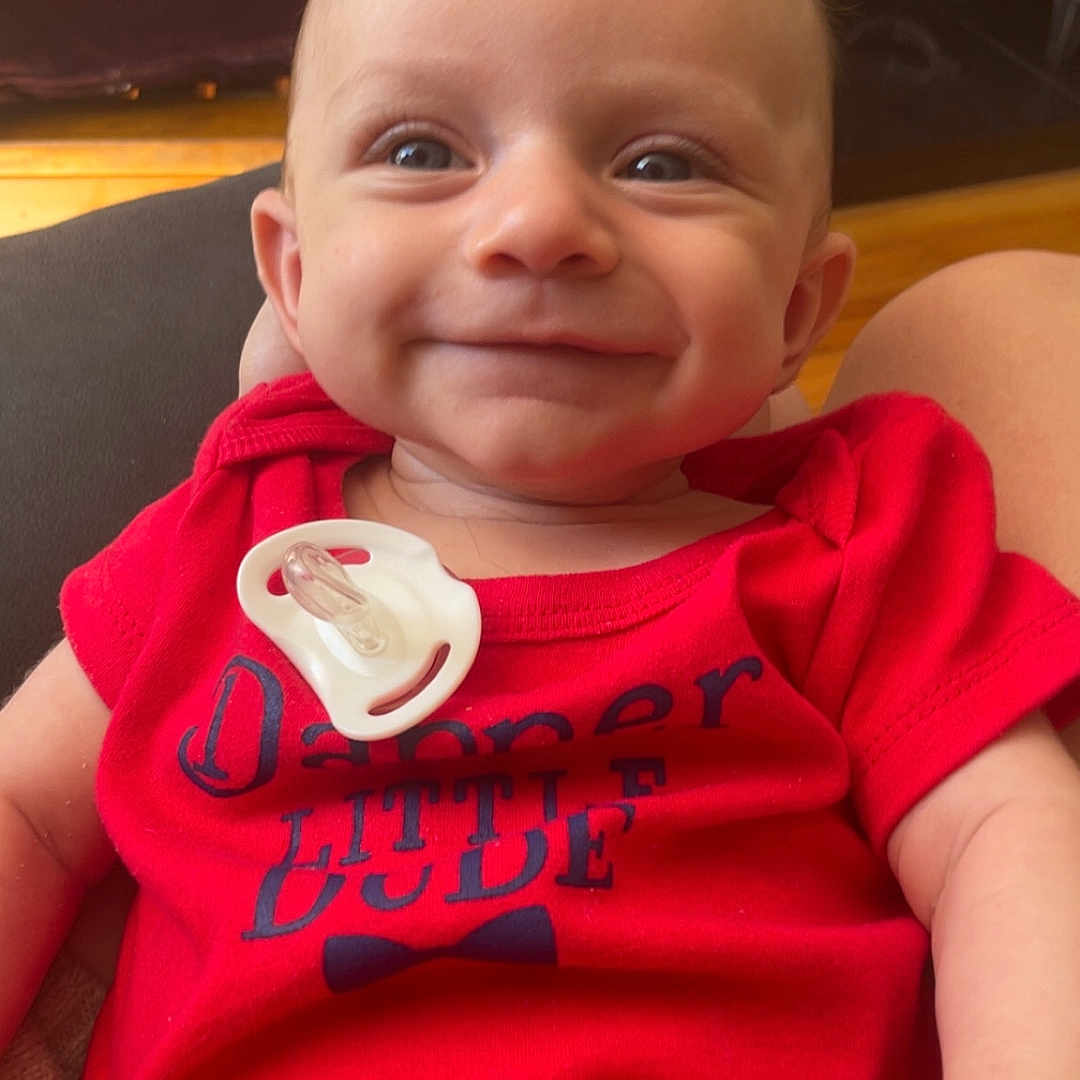 Odin is registered to the contest to win money with this photo: baby, bodypart, clothing, face, finger, happy, head, indoors, laughing, mouth, newborn, pants, person, photography, portrait, shirt, sitting, smile, t_shirt, teeth