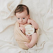 Charles a rejoint le concours — aidez-le/la à gagner de superbes lots ! baby, infant, wrapped, blanket, soft, white, fabric, wooden_sign, holding, curious, wide_eyes, cute, newborn, skin, small_feet, portrait, indoors, peaceful, resting, child