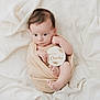 baby, infant, wrapped, blanket, soft, white, fabric, wooden_sign, holding, curious, wide_eyes, cute, newborn, skin, small_feet, portrait, indoors, peaceful, resting, child
