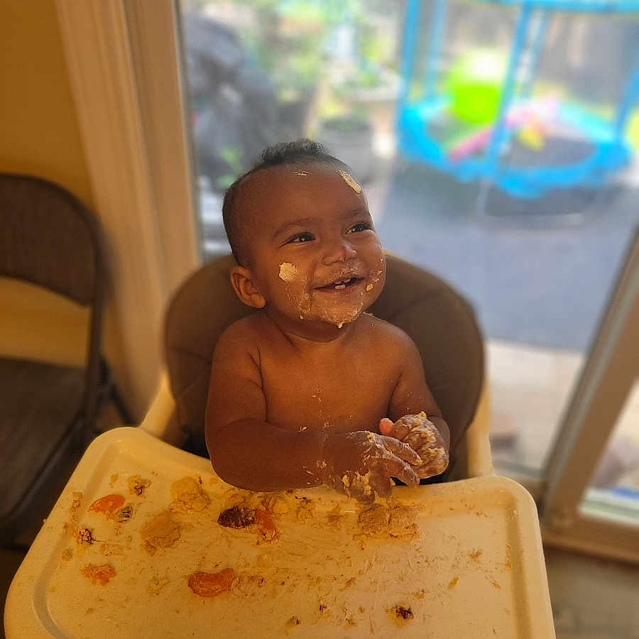 Mateo is registered to the contest to win money with this photo: baby, high_chair, messy, food, smiling, face, hands, indoor, window, play_area, child, happy, tray, skin, mouth, seat, furniture, daylight, expression, young