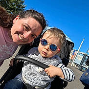 Lucas participe au concours pour gagner de l'argent avec cette photo : child, adult, stroller, sunglasses, smile, outdoor, blue_sky, tree, pavement, person, stroller_handle, jacket, hand, toy, bus_stop, map, reflection, sunlight, family, portrait