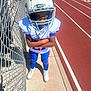child, football_player, helmet, sports_uniform, running_track, chain_link_fence, outdoor, daylight, concrete, shadow, athlete, sport, youth, blue_pants, white_shoes, determined, young_boy, protective_gear, field, team_sports