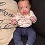 baby, child, curly_hair, denim_pants, floral_shirt, couch, pillow, indoor, sitting, white_socks, cute, portrait, smiling, infant, person, cozy, home, looking_at_camera, clothing, relaxed