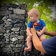 William is registered to the contest to win money with this photo: baby, superman_costume, tattoo, arm, stone_wall, outdoor, greenery, blurred_background, child, person, cute, infant, portrait, holding, skin, short_sleeves, daylight, nature, expression, hands