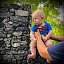 baby, superman_costume, tattoo, arm, stone_wall, outdoor, greenery, blurred_background, child, person, cute, infant, portrait, holding, skin, short_sleeves, daylight, nature, expression, hands
