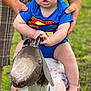 baby, child, superman_onesie, blue_clothing, tattoo, adult_hands, metal_goat, sculpture, outdoor, grass, curious_expression, drool, barefoot, person, infant, playful, summer, cute, nature, fun