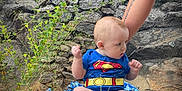 William joined the competition — help win amazing prizes! baby, superman_onesie, blanket, rock, stone_wall, greenery, plant, outdoor, nature, child, tattoo, arm, person, sitting, feet, cute, infant, blue_clothing, support, curious