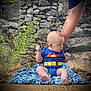 baby, superman_onesie, blanket, rock, stone_wall, greenery, plant, outdoor, nature, child, tattoo, arm, person, sitting, feet, cute, infant, blue_clothing, support, curious