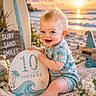 baby, infant, child, beach, sunset, surfboard, sign, smiling, blue_eyes, blonde_hair, knitted_blanket, sand, flowers, ocean, waves, sun, cute, outdoor, portrait, summer