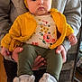 baby, child, infant, yellow_headwrap, yellow_jacket, floral_onesie, green_pants, white_slippers, hands, person_holding, indoor, wooden_floor, gray_sweatshirt, nike_logo, cute, portrait, cozy, soft_slippers, casual_clothing, calm_expression