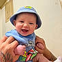 Zara is registered to the contest to win money with this photo: baby, child, smiling, hat, blue_clothing, tattoo, hands, sitting, indoor, cute, infant, happy, person, sock, wall, portrait, skin, face, young, holding