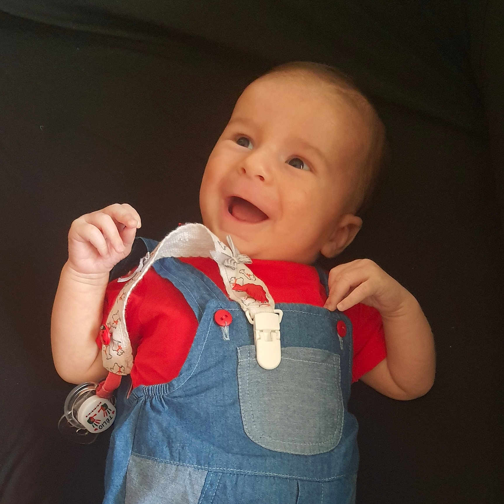 Télio a rejoint le concours — aidez-le/la à gagner de superbes lots ! baby, baby_accessory, child, clothing, cute, denim_overalls, expression, face, hands, happy, head, infant, lying_down, pacifier_clip, playful, portrait, red_shirt, skin, smiling, young