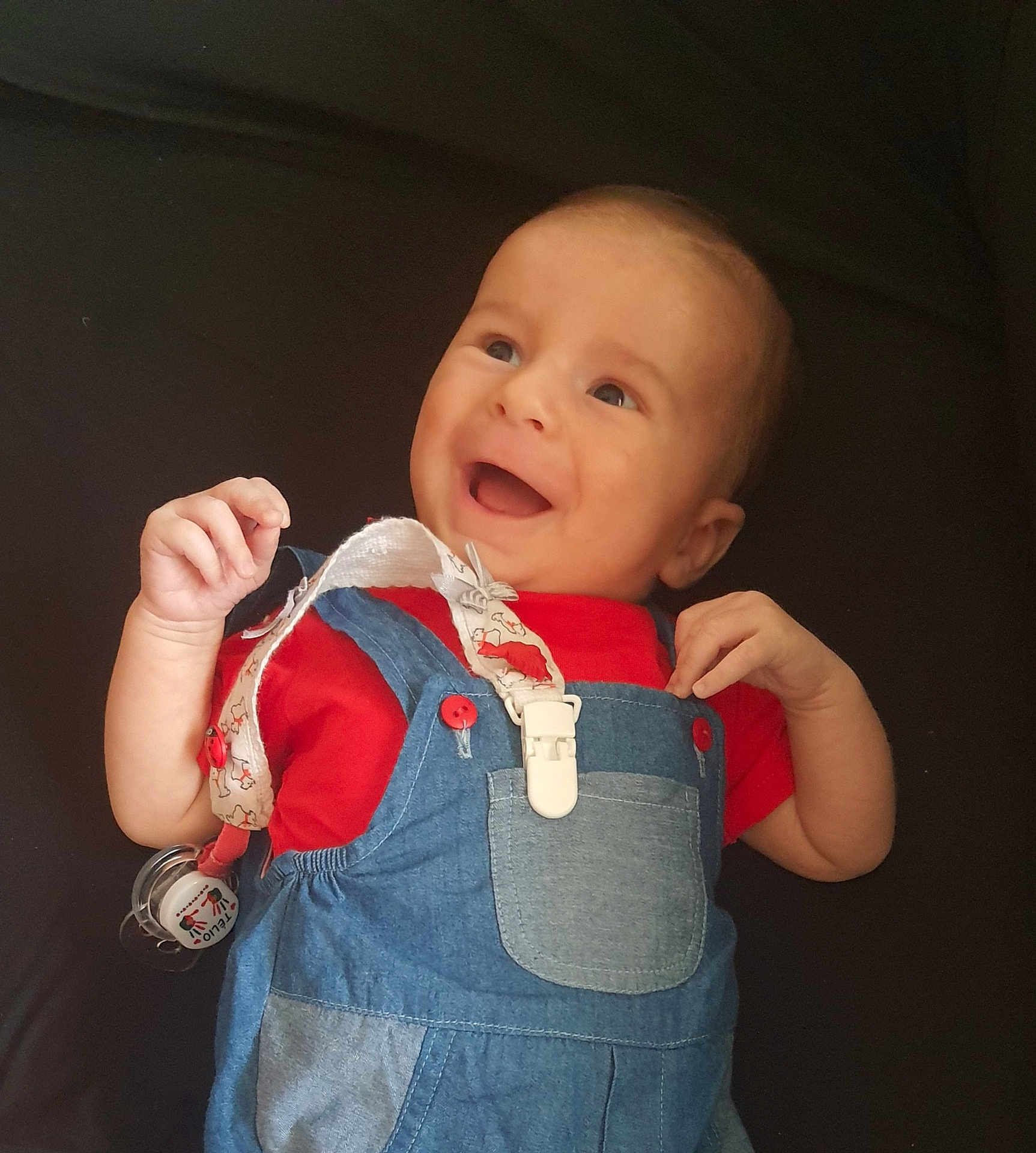 Télio a rejoint le concours — aidez-le/la à gagner de superbes lots ! baby, smiling, red_shirt, denim_overalls, infant, happy, face, hands, clothing, cute, child, portrait, lying_down, playful, young, baby_accessory, pacifier_clip, expression, skin, head
