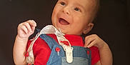 Télio participe au concours pour gagner de l'argent avec cette photo : baby, smiling, red_shirt, denim_overalls, infant, happy, face, hands, clothing, cute, child, portrait, lying_down, playful, young, baby_accessory, pacifier_clip, expression, skin, head