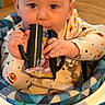 baby, child, infant, high_chair, sitting, indoors, face, portrait, curious, hands, pajamas, patterned_clothing, black_object, wooden_floor, furniture, colorful, close_up, looking_at_camera, home, photography