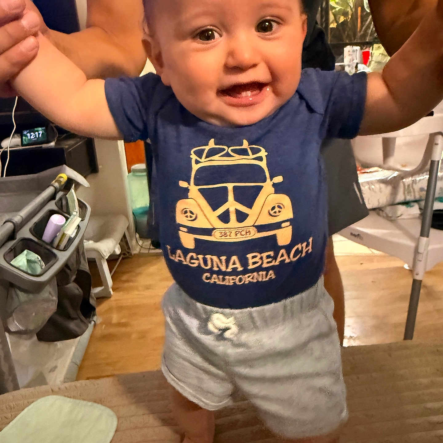 Kasin is registered to the contest to win money with this photo: baby, child, infant, smiling, happy, portrait, indoor, standing, being_held, adult, arms, blue_shirt, graphic_tshirt, shorts, barefoot, baby_gear, stroller, home, wood_floor, family