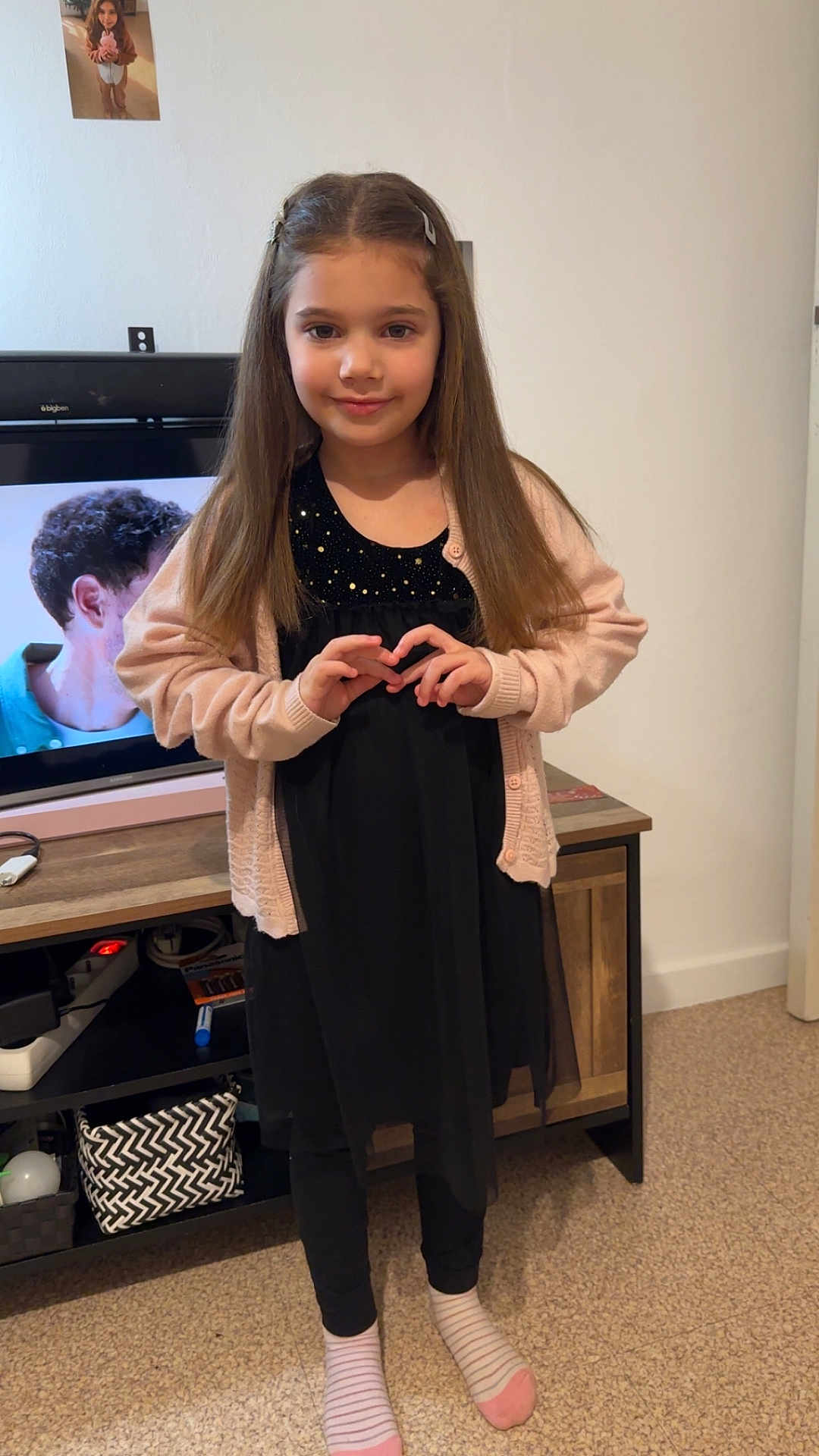 Mia participe au concours pour gagner de l'argent avec cette photo : child, girl, long_hair, cardigan, black_dress, striped_socks, indoor, living_room, television, tv_screen, photo_on_wall, wooden_console, carpet, standing, portrait, smile, hair_clips, heart_hands, furniture, cozy
