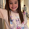 child, girl, smile, pigtails, sweatshirt, cartoon_characters, indoor, hallway, doorway, long_hair, casual_pose, people_in_background, happy, young, childhood, portrait, clothing, bright, person, cute