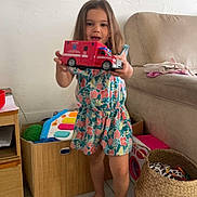 Mia participe au concours pour gagner de l'argent avec cette photo : child, girl, toy_ambulance, floral_dress, living_room, sofa, woven_basket, toy_keyboard, wooden_box, tile_floor, barefoot, smiling, playing, portrait, wall_art, couch, stacked_clothes, toy_bin, colorful_toy, indoor