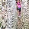 child, rope_bridge, netting, safety_harness, pink_clothing, outdoor, playground, smiling, walking, young_girl, adventure, net, daylight, activity, nature, fun, person, shoes, rope, recreation