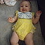 Abby is registered to the contest to win money with this photo: baby, infant, yellow_outfit, smiling, lying_down, blanket, cushion, soft_surface, child, cute, indoors, comfortable, happy, person, clothing, face, head, legs, arm, skin