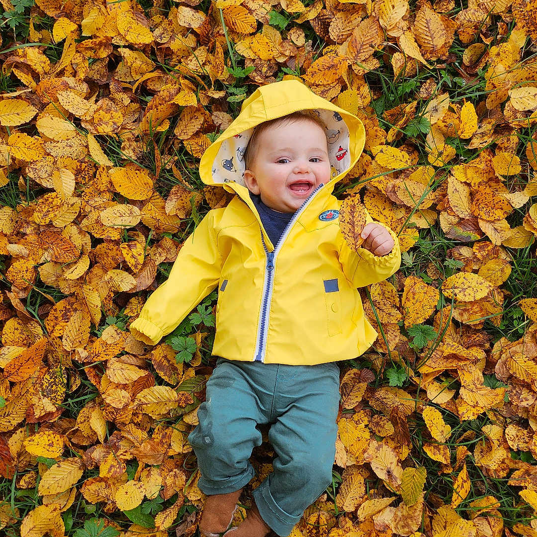 émile a rejoint le concours — aidez-le/la à gagner de superbes lots ! autumn_leaves, baby, baby_clothing, child, cute, fall, fall_colors, grass, green_pants, happy, infant, leaf, nature, outdoor, park, person, playful, seasonal, smiling, yellow_raincoat