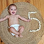 baby, diaper, smiling, happy, infant, floor, wooden_floor, rug, woven_rug, flowers, number_5, milestone, celebration, arms_outstretched, barefoot, indoor, child, cute, portrait, lying_down
