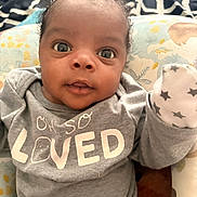 Zaire joined the competition — help win amazing prizes! baby, infant, child, portrait, wide_eyed, lying_down, onesie, mitten, stuffed_toy, cushion, blanket, bed, indoor, close_up, face, eyes, curly_hair, gray_clothing, patterned_background, adorable