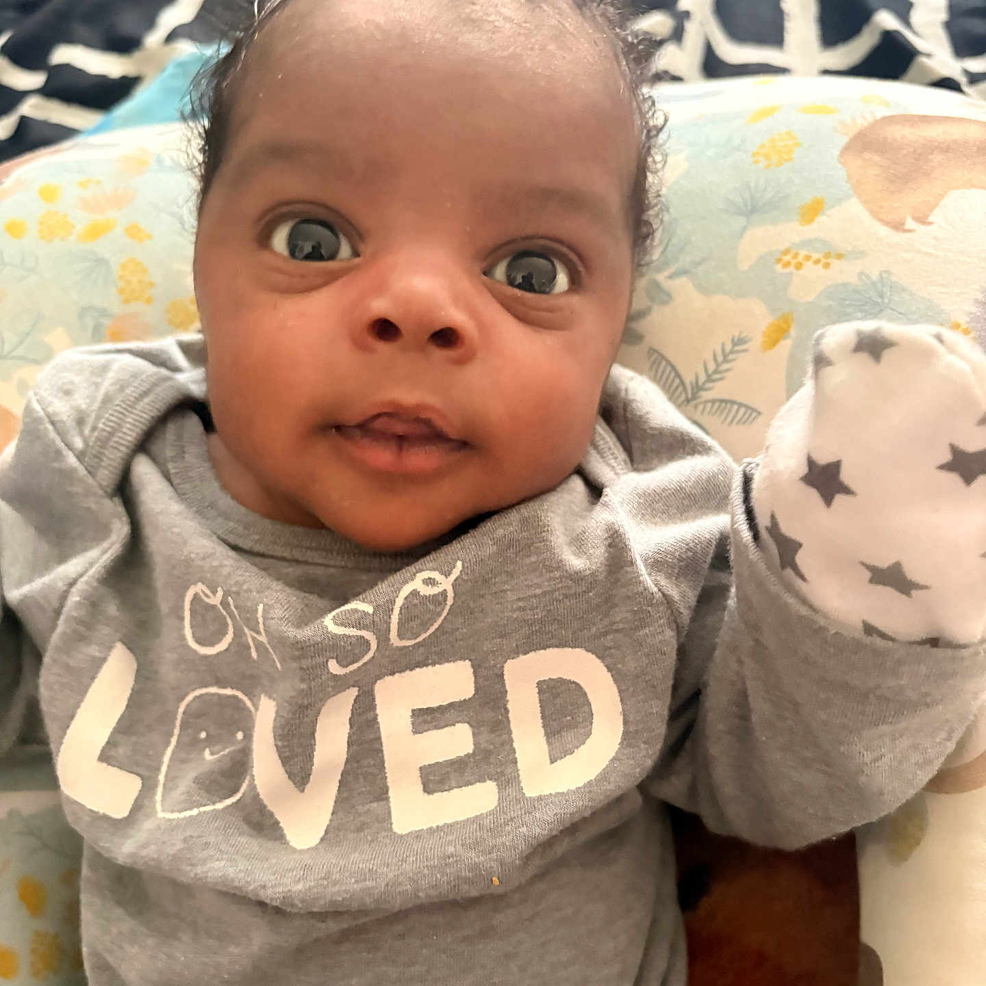 Zaire joined the competition — help win amazing prizes! baby, infant, child, portrait, wide_eyed, lying_down, onesie, mitten, stuffed_toy, cushion, blanket, bed, indoor, close_up, face, eyes, curly_hair, gray_clothing, patterned_background, adorable