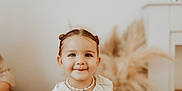 Mia a rejoint le concours — aidez-le/la à gagner de superbes lots ! baby, blurry_background, child, cute, decor, face, hairstyle, happy, indoor, innocent, natural_elements, necklace, person, portrait, shell_necklace, smile, soft_lighting, toddler, white_dress, young_child