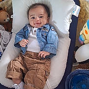 Isaac a rejoint le concours — aidez-le/la à gagner de superbes lots ! baby, infant, smile, denim_jacket, brown_pants, white_shoes, pillow, toys, stuffed_animal, play_area, happy, indoor, child, cute, comfortable, clothing, face, person, cushion, relaxing