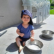 Solann participe au concours pour gagner de l'argent avec cette photo : child, toddler, hat, baseball_cap, blue_shirt, barefoot, metal_bowl, outdoor, patio, tiles, sunlight, shadow, grass, garden, stacked_chairs, smile, playful, person, daytime, casual