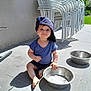 child, toddler, hat, baseball_cap, blue_shirt, barefoot, metal_bowl, outdoor, patio, tiles, sunlight, shadow, grass, garden, stacked_chairs, smile, playful, person, daytime, casual