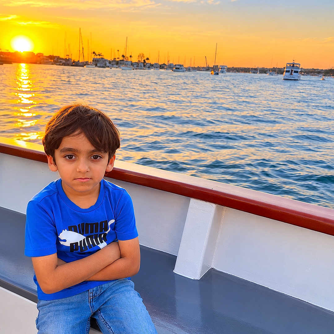 Fahd is registered to the contest to win money with this photo: child, boy, portrait, sitting, boat, sunset, water, marina, harbor, sailboats, ocean, travel, outdoors, blue_shirt, jeans, crossed_arms, serious_expression, sunlight, reflection, seascape