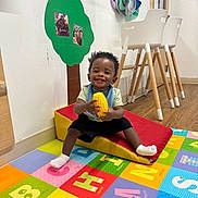 Kayden is registered to the contest to win money with this photo: alphabet, bib, chair, child, daycare, decor, floor, happy, indoor, photograph, play, playmat, shorts, smiling, sock, toddler, toy, wall, wood, yellow