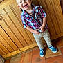 child, smiling, indoor, plaid_shirt, pants, blue_sneakers, wooden_wall, tile_floor, happy, standing, person, short_hair, fashion, casual_clothing, cute, young_child, portrait, holding_object, moawk_hairstyle, light