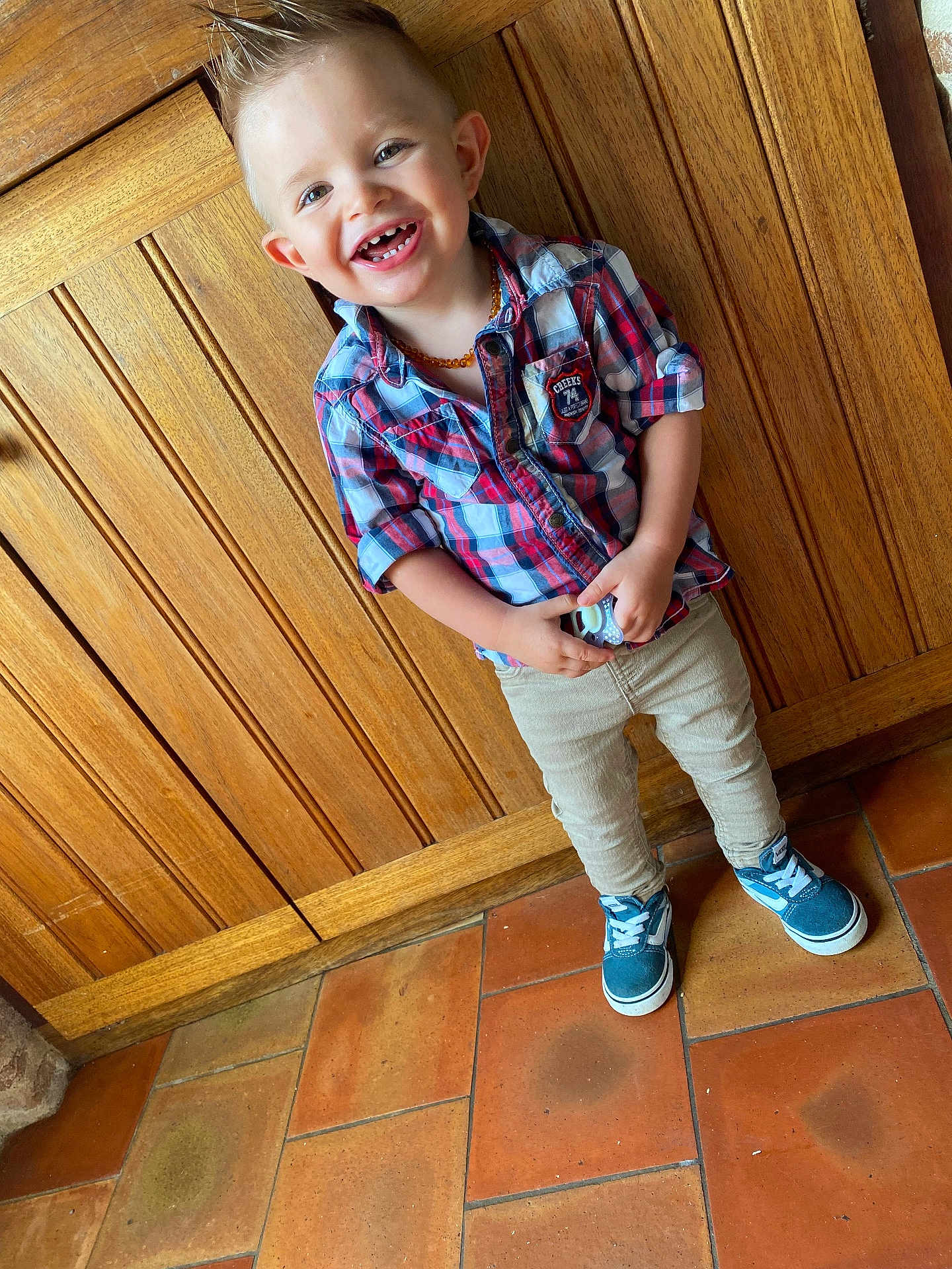 Kaïs a rejoint le concours — aidez-le/la à gagner de superbes lots ! child, smiling, indoor, plaid_shirt, pants, blue_sneakers, wooden_wall, tile_floor, happy, standing, person, short_hair, fashion, casual_clothing, cute, young_child, portrait, holding_object, moawk_hairstyle, light