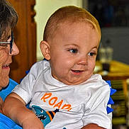 Alistair is registered to the contest to win money with this photo: baby, child, elderly_person, woman, indoor, smile, shirt, glasses, face, person, holding, affection, happy, curious, blue_clothing, white_clothing, short_hair, furniture, background, portrait
