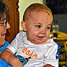 baby, child, elderly_person, woman, indoor, smile, shirt, glasses, face, person, holding, affection, happy, curious, blue_clothing, white_clothing, short_hair, furniture, background, portrait