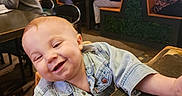 Alistair is registered to the contest to win money with this photo: toddler, child, smile, denim_jacket, table, restaurant, indoor, people, seated, happy, face, cute, wood, casual_clothing, baby, high_chair, background, group, social, light