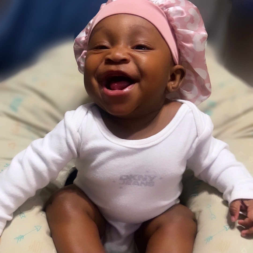 Amyra is registered to the contest to win money with this photo: apparel, baby, baby_sock, child, comfort, cushion, cute, face, happy, headwear, indoor, infant, joy, pink_bonnet, portrait, sitting, smiling, sock, white_onesie, young_child