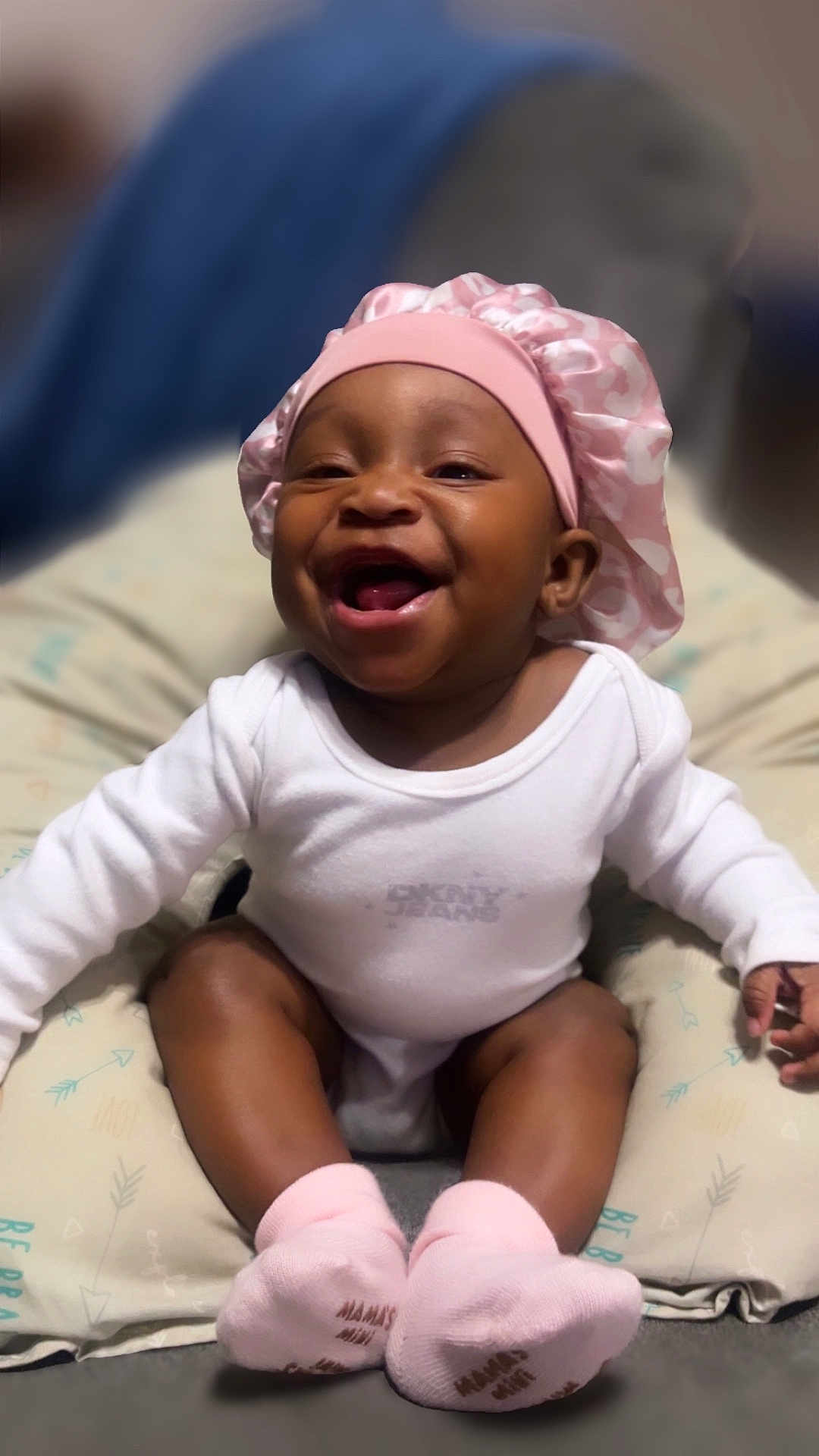 Amyra is registered to the contest to win money with this photo: baby, smiling, pink_bonnet, white_onesie, cushion, sitting, happy, infant, child, cute, sock, baby_sock, indoor, portrait, face, headwear, apparel, comfort, joy, young_child