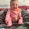 baby, child, pink_clothing, teething_toy, blanket, pillow, bed, smile, happy, indoor, person, infant, cute, cozy, soft_texture, head, hand, sitting, face, nursery